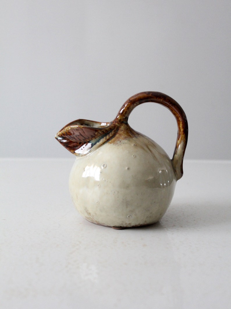 May include: A small, white ceramic pitcher with a brown handle and a brown leaf design. The pitcher has a round body and a small spout.