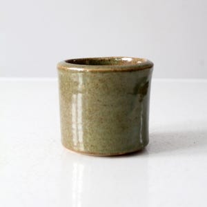 Peut inclure: Petit récipient cylindrique en céramique avec un glaçage vert olive moucheté. Le bord est légèrement évasé et la pièce a un aspect rustique et fait à la main. Le récipient est adapté pour contenir de petits objets.