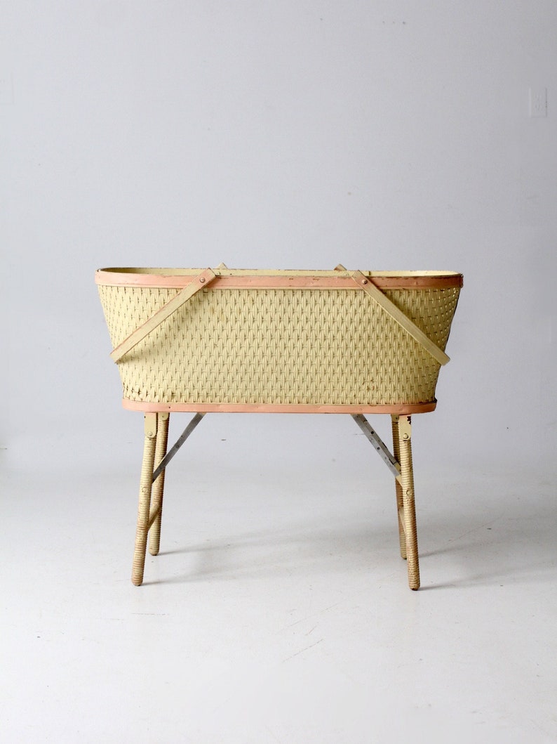 May include: A vintage wicker baby bassinet with a light pink trim and wooden legs. The bassinet is oval shaped and has a woven pattern.