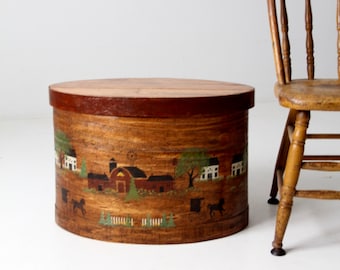 Puede incluir: Una caja de almacenamiento redonda de madera con una escena pintada de una granja y caballos. La caja es marrón con una tapa marrón oscuro y tiene un borde blanco pintado alrededor de la escena.