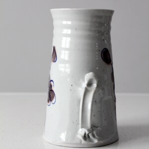 May include: White ceramic pitcher with a brown and blue floral design and a unique handle. The pitcher has a speckled glaze.