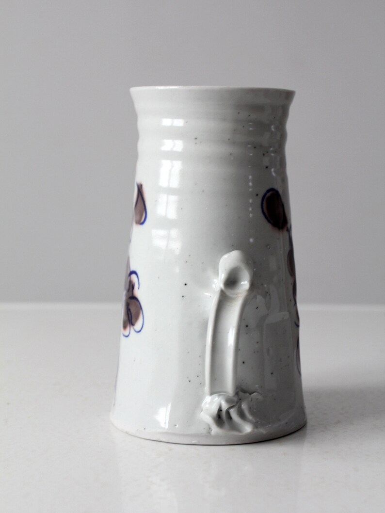 May include: White ceramic pitcher with a brown and blue floral design and a unique handle. The pitcher has a speckled glaze.