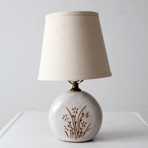 Puede incluir: Una pequeña lámpara de mesa redonda de cerámica con una pantalla de tela beige. La base de la lámpara es de color blanco roto con un diseño floral en marrón y rosa. La lámpara tiene un cuello color latón.