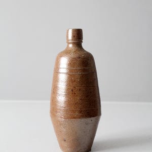 May include: A brown stoneware bottle with a narrow neck and a rounded body. The bottle has horizontal lines around the middle and a two-tone colour scheme, with a lighter base. The bottle is likely an antique or vintage piece.
