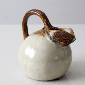 May include: A small, white ceramic pitcher shaped like a pear with a brown handle and a brown leaf detail.