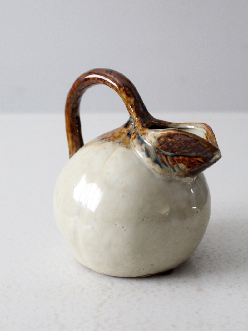 May include: A small, white ceramic pitcher shaped like a pear with a brown handle and a brown leaf detail.