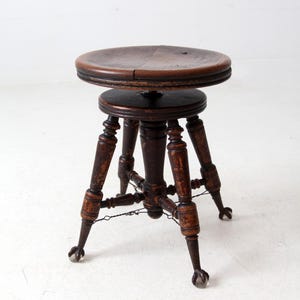May include: Antique wooden piano stool with a round seat and ornate, turned legs. The stool is dark brown with a distressed finish, and features small caster wheels on the feet. A decorative piece of furniture.