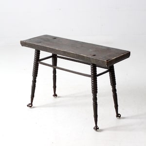 May include: Antique wooden bench with a dark finish. The bench has a rectangular seat supported by turned legs and a connecting stretcher. The legs end in small caster wheels. A vintage piece of furniture.