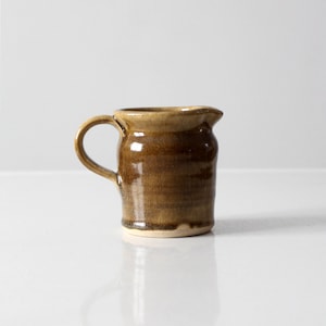 Puede incluir: Una pequeña jarra de cerámica marrón con cuerpo redondeado y asa. La jarra tiene un esmalte marrón claro con un esmalte marrón oscuro en la base.