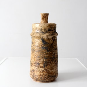 May include: A cylindrical ceramic vessel with a textured, mottled brown and tan glaze. The container features a slightly tapered body, a distinct horizontal ridge near the top, and a small, central knob-like handle on the flat lid.
