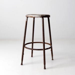 May include: Vintage industrial stool with a round seat and four legs. The seat has a distressed, weathered appearance with remnants of white paint. The legs and footrest are made of rusty metal, giving it a rustic aesthetic.