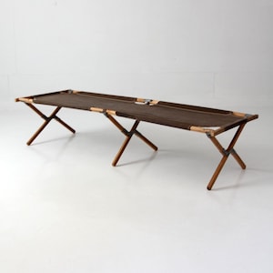 May include: A folding cot with a dark brown canvas sleeping surface stretched across a wooden frame. The structure features three sets of crossed wooden legs connected by metal hinges, allowing the frame to collapse for storage and transport.