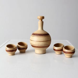 May include: A set of five ceramic drinking vessels, including a decanter and four shot glasses. The decanter has a round body with brown stripes and a wooden stopper. The shot glasses are also round with brown stripes.