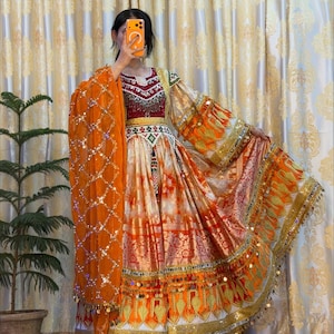May include: A vibrant, traditional dress in shades of orange, red, and gold. The dress features intricate embroidery, beadwork, and embellishments. The long sleeves and flowing skirt are adorned with decorative trim. A matching orange shawl is draped over the shoulder.