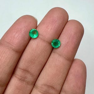 Puede incluir: Dos piedras preciosas de esmeralda verdes redondas y vibrantes. Las gemas se sostienen en una mano sobre un fondo blanco. Las esmeraldas tienen un aspecto brillante y pulido, mostrando su claridad y color. Estas piedras son ideales para la fabricación de joyas.
