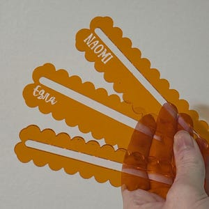 Pode incluir: Três marcadores de página em acrílico laranja translúcido com bordas recortadas e uma fenda vertical central. Cada marcador apresenta um nome personalizado gravado em branco, especificamente NAOMI e Ezra, em uma fonte clara e legível.