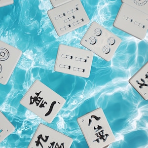 May include: A collection of white rectangular mahjong tiles scattered across a bright blue water surface. The tiles feature various black engraved Chinese characters, circular symbols, and linear patterns on their front faces.