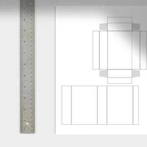 May include: A metal ruler with inch markings is positioned next to a white sheet of paper. The paper features a box template design with fold lines, ideal for crafting or packaging. The ruler is approximately 28 cm long.