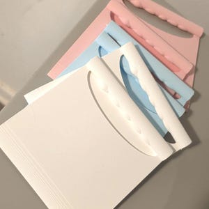 May include: A stack of three square plastic scrapers, each featuring a curved handle with a scalloped grip. The top scraper is white, the middle one is light blue, and the bottom scraper is light pink. Each tool has a flat, straight edge.