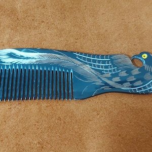 Puede incluir: Un peine azul oscuro con un diseño decorativo de pájaro. El peine presenta un tallado detallado de un pájaro con detalles de plumas blancas y un ojo amarillo. El peine está sobre una superficie marrón.