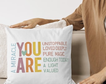 May include: White square pillow with the words "YOU ARE" in colorful embroidered letters. The words "A MIRACLE" and inspirational phrases like "UNSTOPPABLE" and "PURE MAGIC" are also embroidered on the pillow.