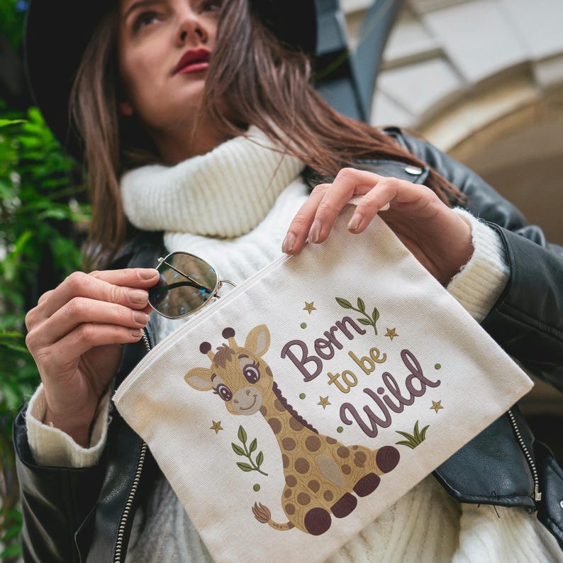 May include: A cream-colored canvas zipper pouch featuring a cartoon giraffe illustration and the text Born to be Wild in dark brown lettering, accented with small gold stars and green leaf motifs on the front surface of the fabric accessory.