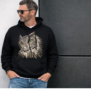 May include: Black hooded sweatshirt featuring a detailed owl graphic on the front. The owl design incorporates shades of brown, gray, and green, with a focus on the bird's face. The sweatshirt has a kangaroo pocket and a drawstring hood.