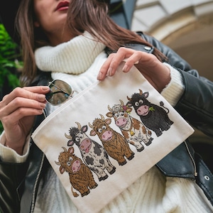 May include: A cream-colored canvas zipper pouch featuring an embroidered graphic of five cartoon cows in a row. The cows vary in color, including brown, black and white, and tan, with one wearing a flower and a bell. The pouch has a simple rectangular shape.