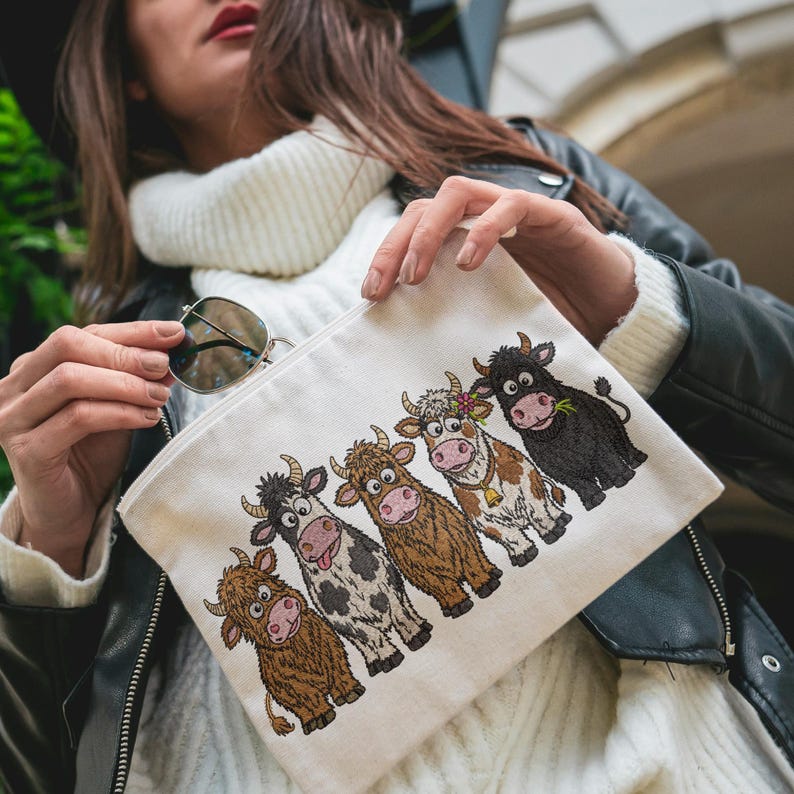May include: A cream-colored canvas zipper pouch featuring an embroidered graphic of five cartoon cows in a row. The cows vary in color, including brown, black and white, and tan, with one wearing a flower and a bell. The pouch has a simple rectangular shape.
