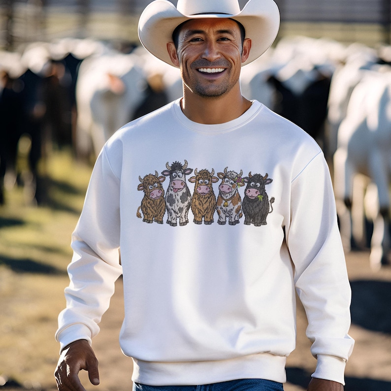May include: A cream-colored crewneck sweatshirt featuring a graphic print of five cartoon cows standing in a row. The cows vary in color, including brown, black and white, and tan, with one wearing a small bell. The sweatshirt has long sleeves.
