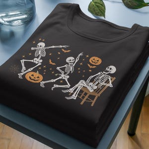 May include: Black t-shirt featuring a Halloween-themed design. The graphic includes three white skeletons in various poses, pumpkins, bats, and stars. One skeleton is dabbing, another is dancing, and the third is relaxing in a chair.