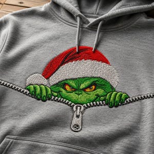 May include: A heather gray hooded sweatshirt featuring an embroidered graphic of a green character with yellow eyes wearing a red and white Santa hat. The design depicts the character peeking out from behind a horizontal silver zipper detail.