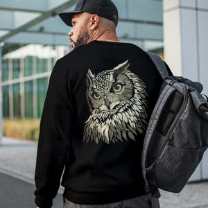 May include: Black sweatshirt featuring a detailed owl graphic on the back. The owl has green eyes and a detailed feather design in shades of gray and white. The sweatshirt is worn with a black cap and a gray backpack.