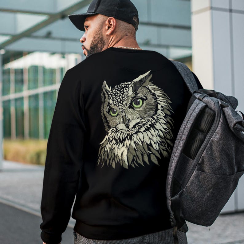 May include: Black sweatshirt featuring a detailed owl graphic on the back. The owl has green eyes and a detailed feather design in shades of gray and white. The sweatshirt is worn with a black cap and a gray backpack.