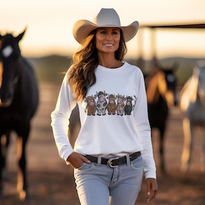 May include: A white long-sleeve sweatshirt featuring a graphic print of five cartoon cows in various colors and patterns, worn with light blue denim jeans and a dark belt. The sweatshirt is paired with a light-colored cowboy hat.