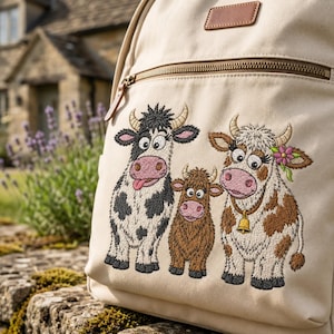 Könnte beinhalten: Ein beigefarbener Rucksack aus Segeltuch mit einer Stickerei von drei Cartoon-Kühen. Die linke Kuh ist schwarz-weiß mit herausgestreckter Zunge, in der Mitte ein kleines braunes Kalb und rechts eine braun-weiße Kuh mit gelber Glocke und Blume.