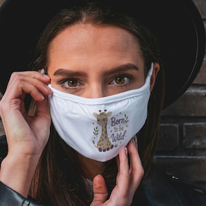 May include: A white face mask featuring a graphic illustration of a brown and tan giraffe. The design includes green leaf accents, small gold stars, and the text "Born to be Wild" printed in a dark brown, stylized script font on the front.