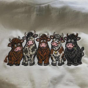 May include: A white sweatshirt featuring an embroidered design of five cartoon cows standing in a row. The cows vary in color, including brown, spotted gray, dark brown, white and brown with a bell, and solid black, all with pink noses.