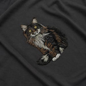 Könnte beinhalten: Gesticktes Design einer langhaarigen Katze mit braunem, schwarzem und weißem Fell und gelben Augen. Die Katze liegt auf einem dunkelgrauen Stoff. Die Stickerei ist detailliert und strukturiert und erzeugt eine realistische Darstellung der Katze.