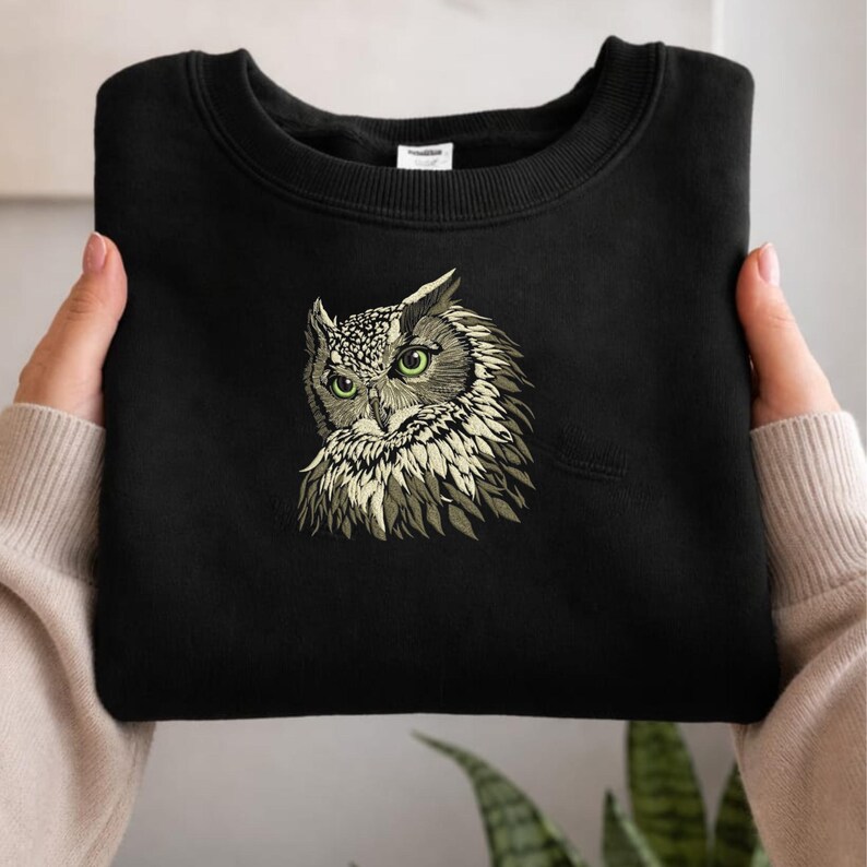 May include: Black sweatshirt featuring an embroidered owl design. The owl has green eyes and detailed feathering in shades of beige and gray. The sweatshirt is folded, showcasing the owl's face prominently.