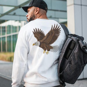 May include: A white long-sleeve sweatshirt featuring a detailed graphic of a bald eagle in flight on the back. The eagle has brown feathers, a white head and tail, and yellow talons. The sweatshirt is worn with a black baseball cap and a dark gray backpack.