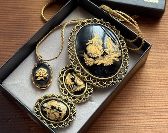 May include: A vintage jewelry set featuring a necklace, earrings, and a brooch, all with black backgrounds and gold-colored filigree frames. The necklace has an oval pendant with a gold-colored design. The earrings and brooch have similar designs.