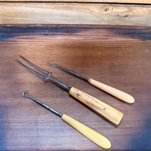 May include: Three antique crochet hooks and a fork-like tool with ivory-colored handles and metal shafts. The tools are arranged on a wooden surface, showcasing their vintage design and craftsmanship.