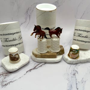 May include: A collection of decorative thimbles, including two with miniature horse figurines and one with a swan design. The thimbles are displayed on small, white stone platforms, with packaging tubes in the background.