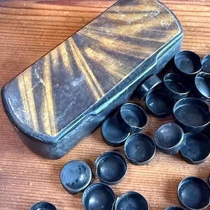 May include: A rectangular, antique wooden box with a lid featuring a sunburst design in shades of brown and gold. Scattered around the box are numerous small, dark blue metal caps. The items are arranged on a wooden surface.