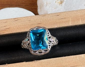 May include: A silver ring with a large, rectangular, bright blue gemstone. The ring has an ornate, filigree design around the gemstone and on the band. The ring is displayed in a wooden jewelry box.