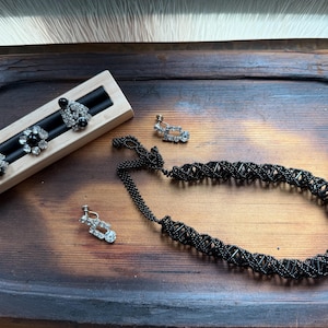 May include: A collection of black jewelry displayed on a wooden tray. The set includes a beaded necklace, two pairs of earrings, and three decorative earrings on a wooden holder. The earrings feature rhinestones and black accents.