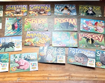 May include: A collection of colorful Beanie Baby trading cards, each featuring a different animal character with a unique name. The cards are arranged on a wooden surface, showcasing various designs and illustrations.