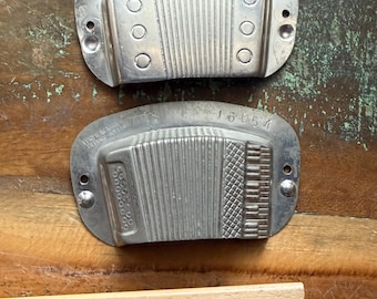 May include: Two silver-toned metal accordion-shaped plates with the text 16054 and VORRENAAR TILBURG-HOLLAND stamped on them. The plates feature vertical ridges and circular indentations. A wooden ruler is positioned below, showing a width of 4 inches.