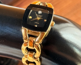 May include: Gold-toned Armitron watch with a black rectangular face and a small diamond accent. The watch features the word "Now" on the face and a linked bracelet. The watch is displayed on a black surface.